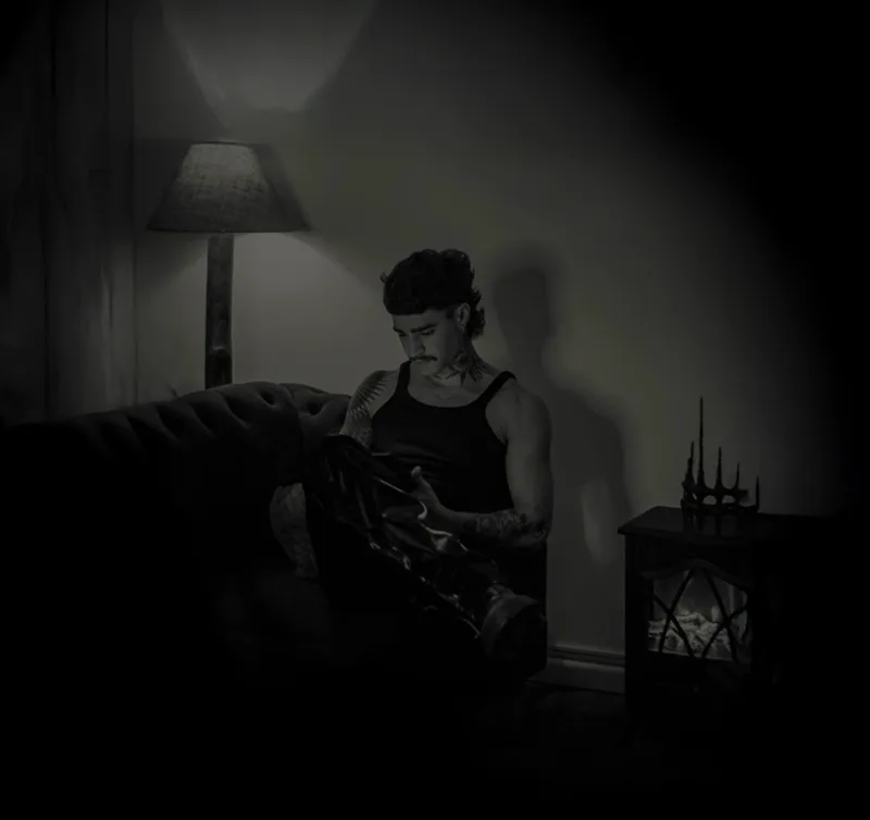 Mateus sentado no sofá do estúdio, à luz de um abajur, preparando o material de tatuagem.