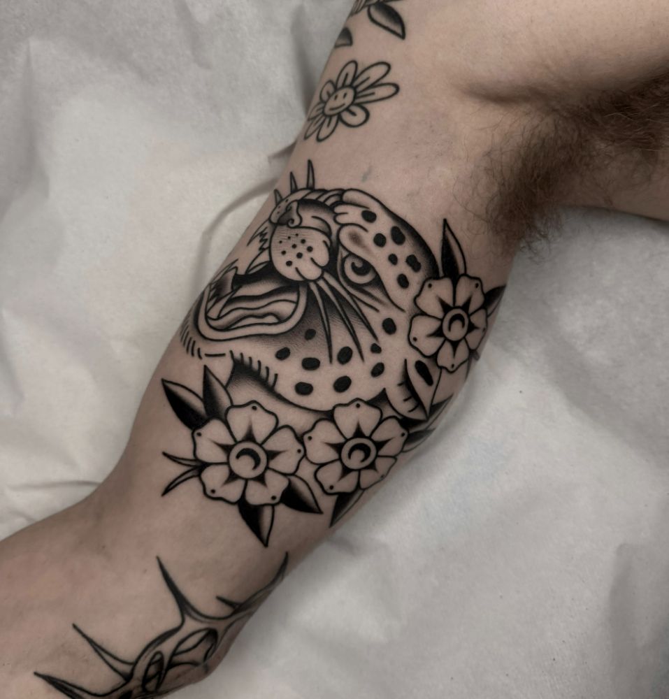 Tatuagem de onça pintada cercada de flores, no braço.